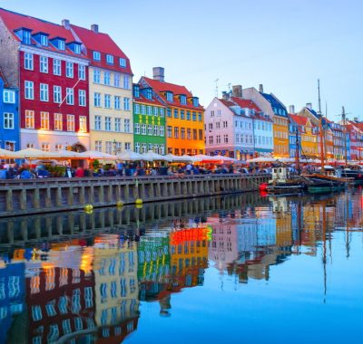 People walking and sitting in restaurants at illuminated Nyhavn embankment by canal with moored boats in evening dusk, Copenhavn, Denmark