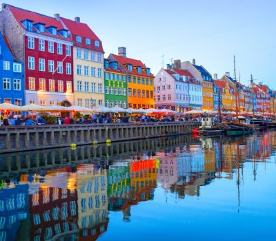 DenmarkCopenhagenNyhavn People walking and sitting in restaurants at illuminated Nyhavn embankment by canal with moored boats in evening dusk, Copenhavn, Denmark